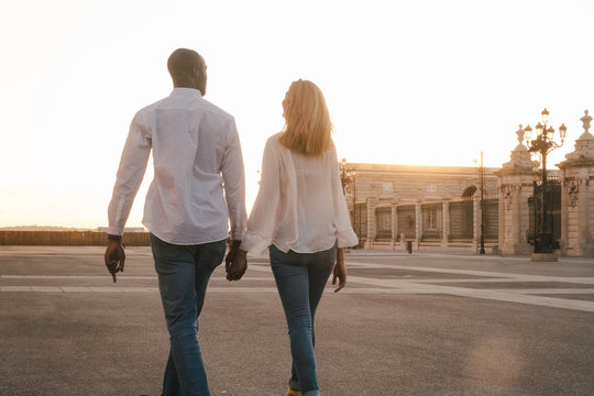 Back view anonymous diverse couple in casual outfits walking together along asphalt sidewalk on street during sunset time