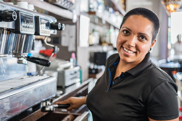 Side view of positive ethnic barista pouring hot coffee into cup from coffee machine while working in bar