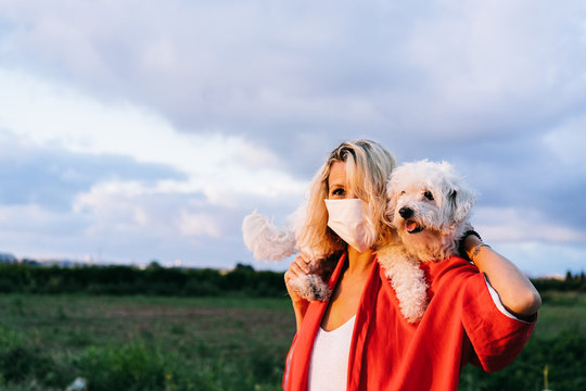 Positive female in casual clothes wearing protective face mask standing with funny white Bichon Frise dog on shoulders against blurred summer countryside landscape during coronavirus outbreak