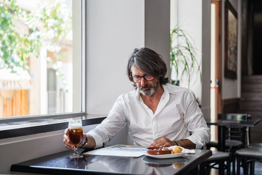 Positive Ethnic Bearded Aged Man Sitting At Table With Of Glass Of Foamy Beer And Reading Newspaper Near Window