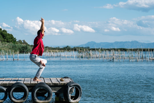 Side view of unrecognizable male in casual clothes performing Eka Pada Utkatasana posture during morning yoga session on wooden quay against calm sea