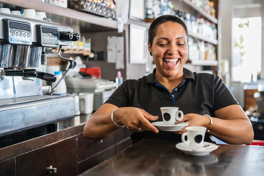 Smiling ethnic female worker in black uniform standing at bar with cups of coffee at daytime