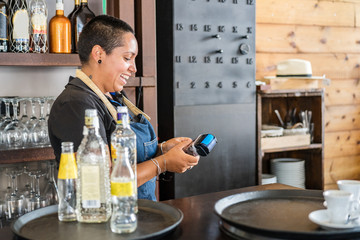 Positive female barkeeper in apron standing at counter with alcohol drinks and smiling while charging costumer on POS machine and credit card working in cafe