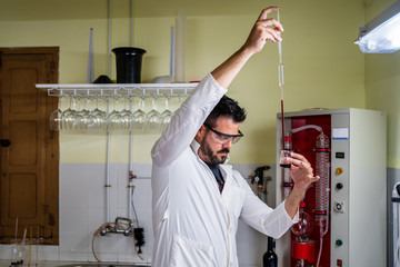 Side view of bearded male chemist in white robe pouring red liquid into transparent flask from long tube while standing near distillation equipment in lab