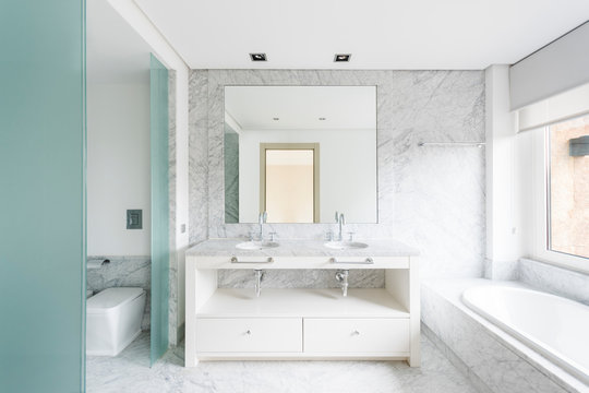 Creative minimalist design of bathroom with similar vanity units under big mirror and bath tube near window