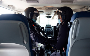 Policemen in medical mask and protective gear talking on radio set while sitting in car with partner
