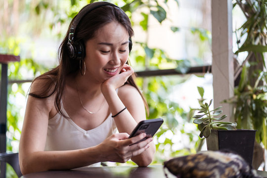 Young ethnic female in summer outfit sitting in headphones at table and enjoying funny video on social media via cellphone