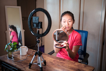 Young Asian woman in casual wear holding mirror and applying foundation on face while recording video tutorial for beauty blog at home workplace with ring light and camera