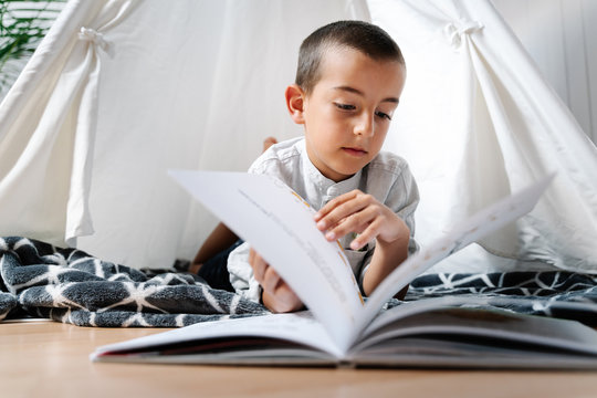 Full body little boy sitting on blanket on wooden floor while reading book in homemade tent decorated with garland of flags in apartment in daylight