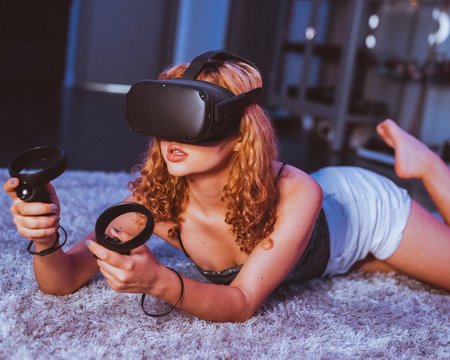 Seductive barefoot anonymous female in casual wear lying with open mouth on soft plaid on bed while playing game with controllers and in goggles of virtual reality