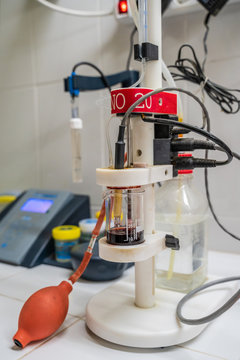 Modern apparatus representing alcohol acidity meter with connected cables and metal part on edge immersed into transparent flask near plastic bottle and ceramic wall in lab
