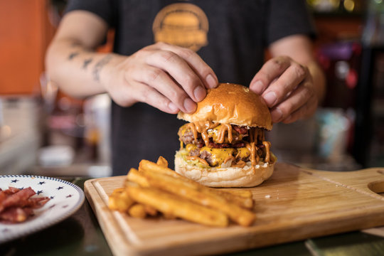 Crop ethnic male cook standing at counter in cafe with delicious burger on chopping board