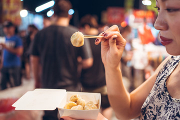 Side view of crop ethnic female standing on crowded street in Zhubei Night Market and eating delicious fried dumplings from paper container