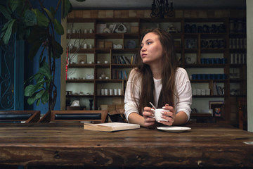 Pensive young woman with long hair relaxing at wooden table with book and drink coffee in cozy cafe