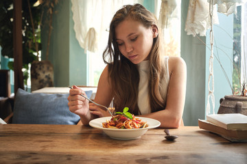 Upset young female relaxing at wooden table with served plate of delicious pasta in cozy restaurant on sunny day