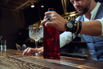 Cropped professional young bartender holding red bottle while preparing a cocktail in the bar