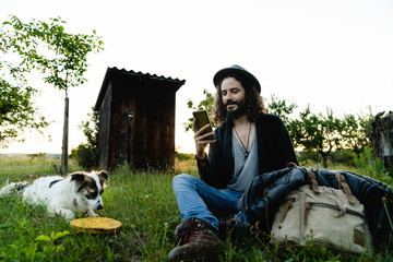 Delighted male hipster sitting on green filed in summer next to a dog and chatting on social media during weekend