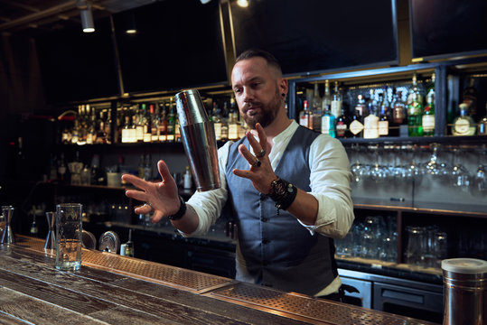 Happy professional barman in elegant suit mixing cocktail in shaker while working in bar looking away