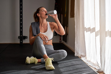 Full body of slim lady drinking fresh water while resting on floor near window during fitness workout