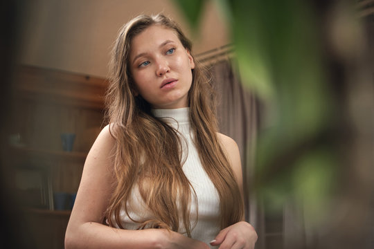 Thoughtful young slim lady with long hair in mini skirt and top standing in front of green plants and looking down