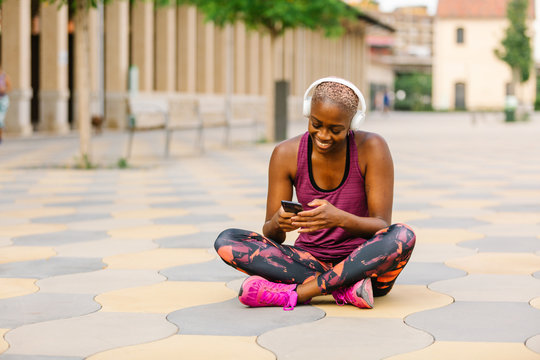 Positive African American female athlete in sports clothes and headphones sitting leaned forward with crossed legs on embankment near building in town browsing smartphone while listening to music