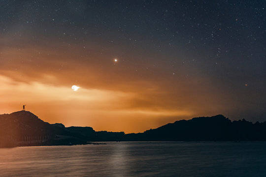 Silhouette of man pointing finger the stars while observing the night sky with the Moon and the stars in the background near to lake