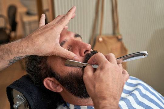 Crop anonymous tattooed barber doing beard hairstyling with sharp metal razor in barbershop