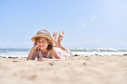 Ground level of full body of cute little boy propping chin while chilling on sandy coast during vacation