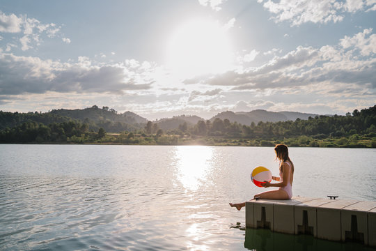 Side view of woman in bikini playing with beach ball while sitting in the edge of wooden pier near pond during summer holiday