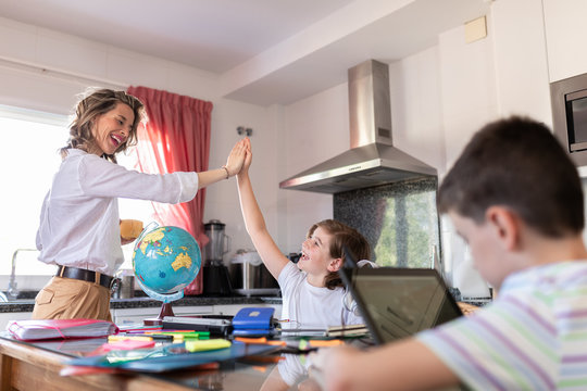 Cheerful tutor in formal wear giving high five to excited child while sitting at table with globe near anonymous boy with laptop during geography lesson at home