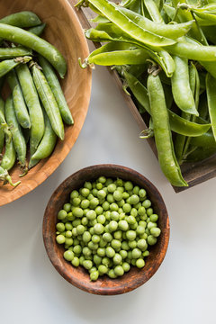 From above of wooden bowl with crispy green peas near pile of crunchy pea pods with spiky edges on white table at home