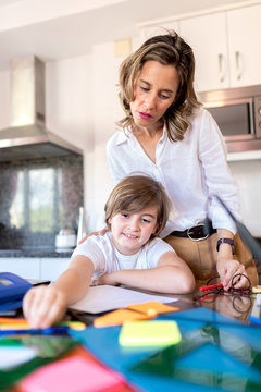 Serious adult woman in formal outfit revising homework standing near smiling boy sitting at table with multicolored stickers while studying in kitchen
