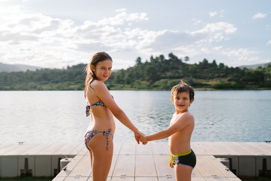 Side view of smiling brother and sister in swimwear standing together holding hands on quay near lake and looking at camera while enjoying summer vacation