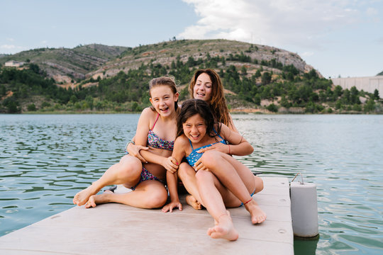 Adult woman in swimwear tickling cheerful children while sitting relaxing on pier embracing each other during summer holiday