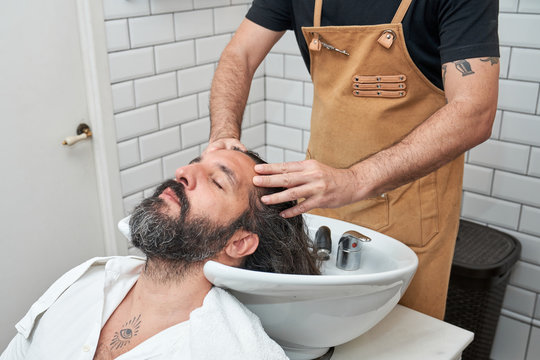 From above crop male barber washing head of bearded relaxing male customer in sink in barbershop