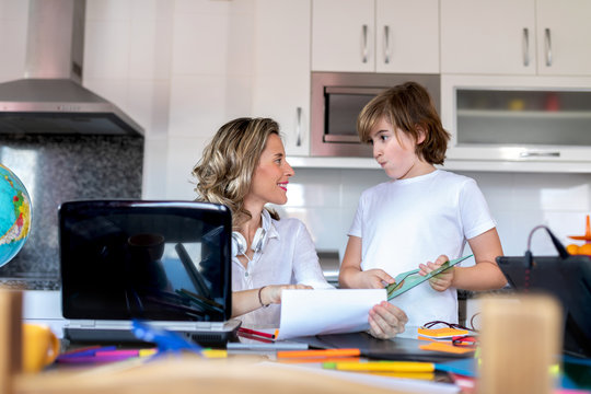 Smiling mother doing homework with boy while looking at each other and sitting at table near netbook with paper and colorful stationery in kitchen