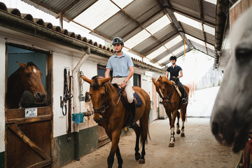 Serious female jockeys in helmets and boots riding chestnut horses along barn on ranch and looking away