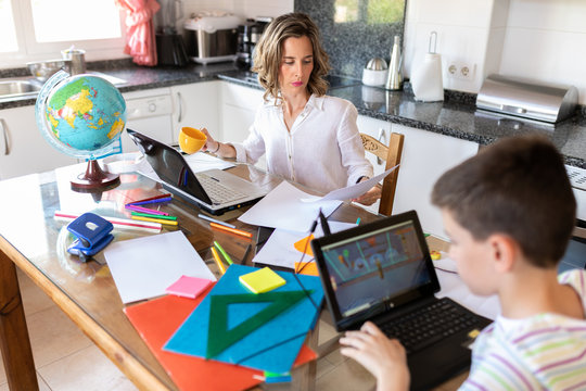 Unrecognizable boy doing homework while typing on netbook and sitting at table near positive mother with blank papers speaking with friend during video chat in kitchen