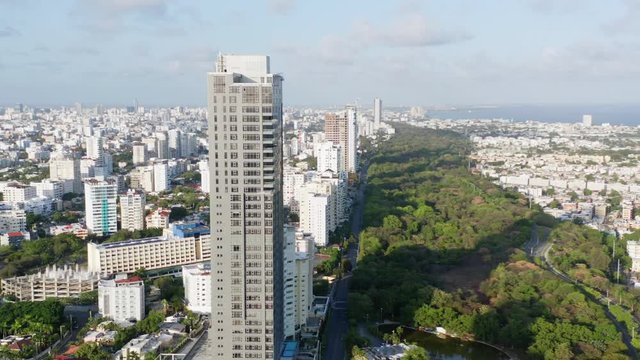 Scenic city skyline aerial view of tall residential Avenida Anacaona skyscraper and downtown Santo Domingo center and view of ocean sea water on sunny day, Dominican Republic, overhead drone descend