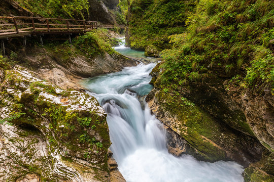 Wide angle view of the Slovenian gorge of Vintgar, with a torrent running between rocks and into rapids surrounded by vegetation, with a wooden walkway on the left