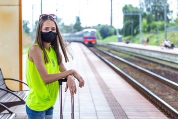 Safe travel. Caucasian teenager in black face mask with suitcase is waiting for train at the railway station.