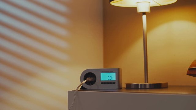Moving shot of a device on a sideboard messuring the power consumtions when a light is turned on. Low energy consumtion of a LED smart home lamp while the lamp changes from warm to cold.