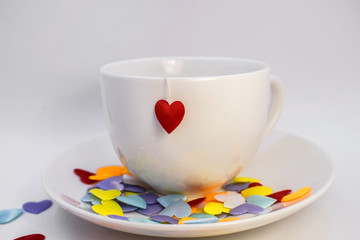 on a light background stands a white Cup on a saucer with a heart hanging from it. On the saucer are colored scattered hearts