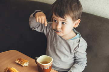 Upper photo of small caucasian brunette boy drinking a tea with spoon at the table and eating some cookies