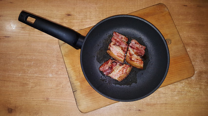 photo of pieces of bacon on a black frying pan on a wooden cutting board on a wooden table