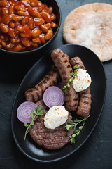 Grilled balkan meat patty or pljeskavica with cevapcici or skinless beef sausages, prebranac and pita, vertical shot over black stone surface