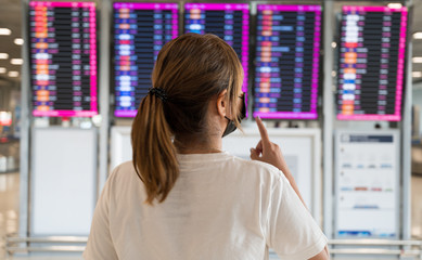 Beautiful young tourist girl  wearing face mask looking airline flight status near flight information board in airport during  covid-19 virus outbreak a new normal concept