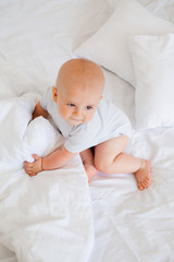 Cute baby boy 6 months smiling in a white bodysuit sitting on a bed on white bedding