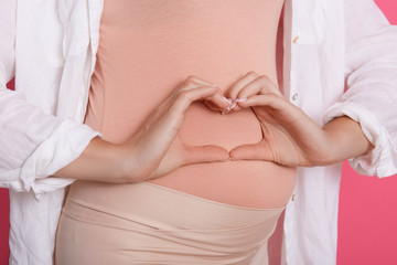 Faceless pregnant woman wearing beige outfit posing with heart symbol against her belly, expressing great love, unknown expectant mother in white shirt.