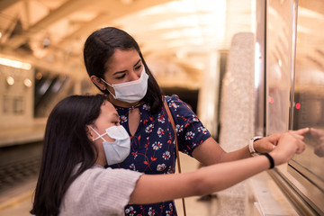 Two latin sisters pointing the poster on the train station wearing disposable mask to prevent coronavirus contagion.